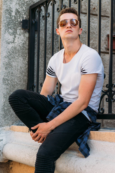 Attractive, sexy guy sits leaning on a curb on a ledge in the wall on the street in sunglasses. self-confidence, flatulence and arrogance.model, posing, portrait of a young man in white sneakers - Photo, Image