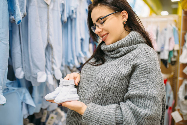 Щасливе майбутнє мати купує дитячий одяг в магазині для новонароджених. Вагітна жінка в магазині товарів для немовлят
 - Фото, зображення