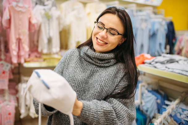 Lächelnde zukünftige Mutter wählt Baby-Hut im Laden für Neugeborene. Schwangere in Geschäft mit Waren für Säuglinge - Foto, Bild