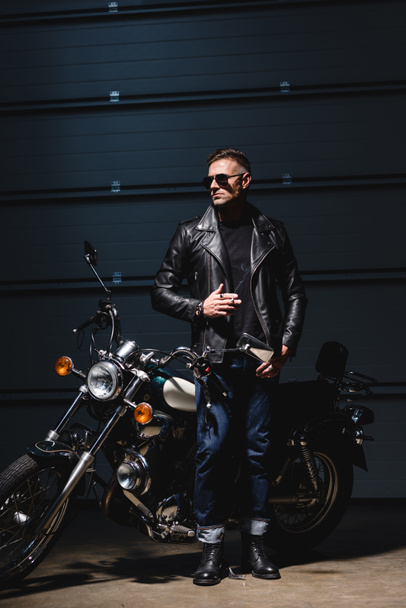 classic guy in sunglasses standing by motorcycle in garage and smoking cigarette - Fotó, kép
