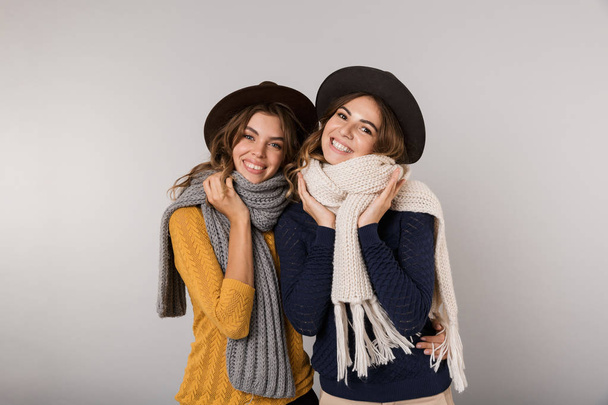 Изображение двух довольных девушек в шляпах и шарфах, улыбающихся в камеру на сером фоне
 - Фото, изображение