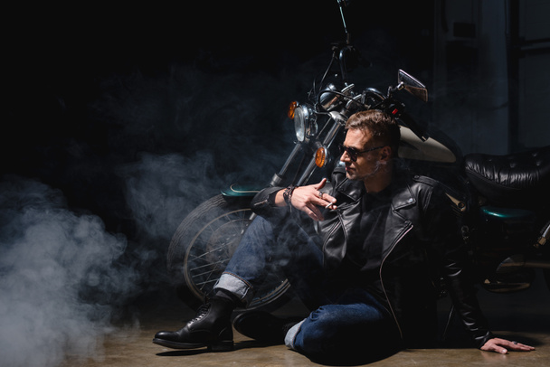 knappe biker in zwarte zonnebril zittend op de vloer vervolgens naar motorfiets in garage - Foto, afbeelding