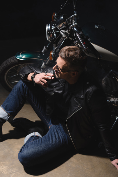 handsome man in black sunglasses and leather jacket sitting on floor next to motorcycle - Foto, imagen