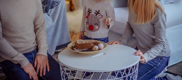 the family sits at the table and makes crafts, is going to decorate the house, on the table are wooden bars, a wooden cut, a log house, a Christmas tree toy,cones. - 写真・画像