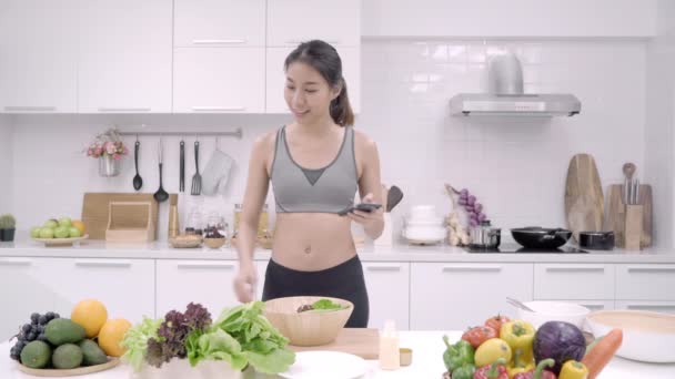 Mujer asiática joven haciendo ensalada comida saludable mientras usa el teléfono móvil para buscar receta en la cocina, hermosas verduras de uso femenino preparando ensalada para el cuerpo en forma en casa. Concepto de comida saludable
. - Imágenes, Vídeo
