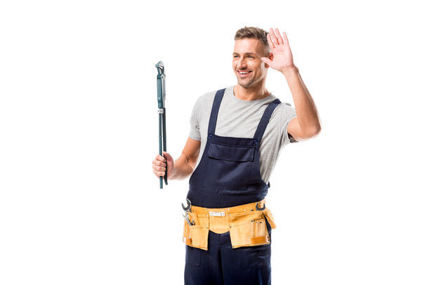 trabajador feliz sosteniendo la llave de tubo, saludando y mirando hacia otro lado aislado en blanco
 - Foto, Imagen