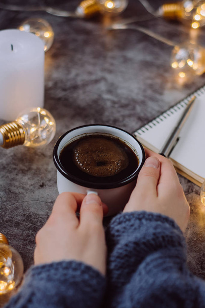 Hand holds  cup of coffee on gray table with notebook, pen and  candle decorated with led lights.Winter and Autumn weekend concept. Flat lay, top view. - Φωτογραφία, εικόνα