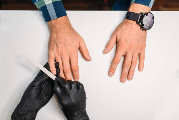 Female master in black gloves polishing nails to male client, top view, men manicure in salon. Manicurist doing hands care procedure - Foto, immagini