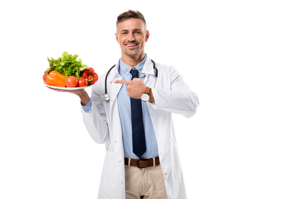 sonriente médico señalando con el dedo el plato de verduras frescas y vegetación aislada en blanco, concepto de alimentación saludable
 - Foto, Imagen