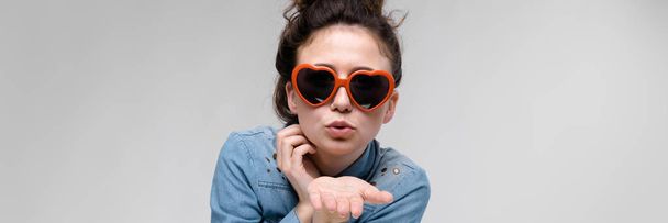 Young brunette girl with glasses in the form of a heart. The hairs are gathered in a bun. The girl sends an air kiss. - Foto, imagen