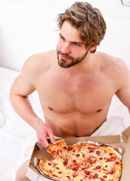 Portrait of lazy muscular man eating pizza while laying on a bed at home. Cropped image of shirtless sexy man with pizza on bed. Break diet concept. - Foto, Imagem