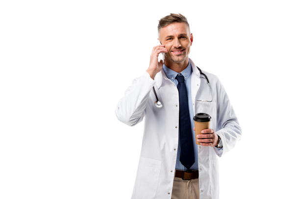doctor looking at camera, holding coffee to go and talking on smartphone isolated on white - Photo, Image