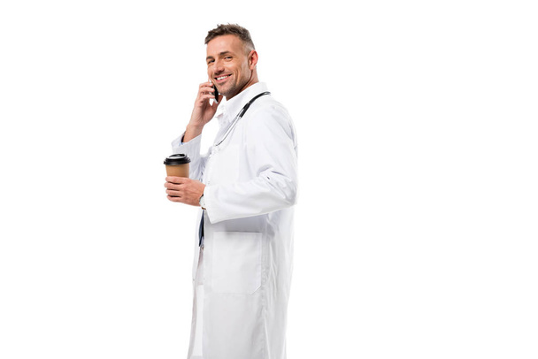medico sorridente guardando la fotocamera, tenendo il caffè per andare e parlando su smartphone isolato su bianco
 - Foto, immagini