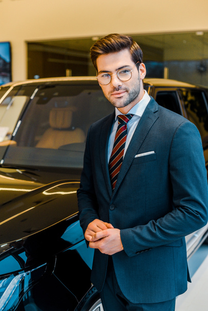 happy young businessman in eyeglasses posing near car in showroom - Foto, Imagem