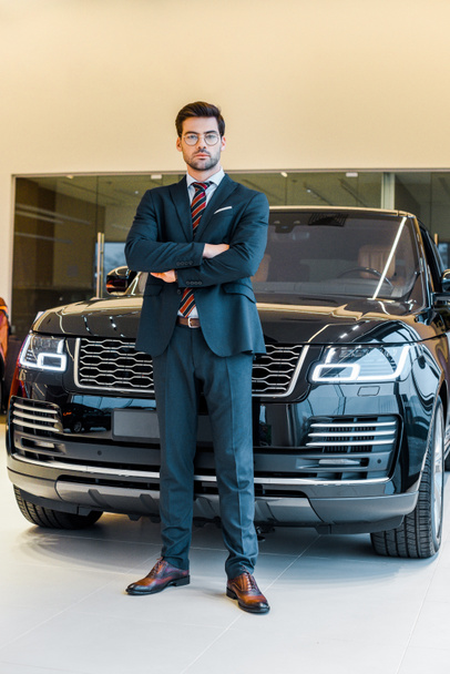 low angle view of businessman in eyeglasses posing with crossed arms near black automobile - Φωτογραφία, εικόνα