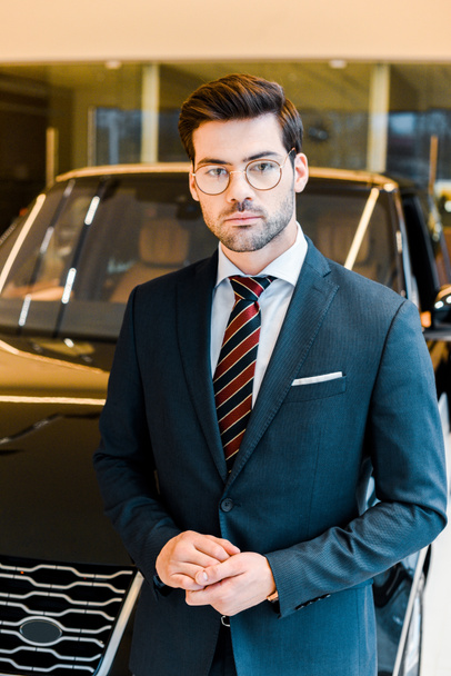 selective focus of young businessman in eyeglasses looking at camera near car in showroom - Valokuva, kuva
