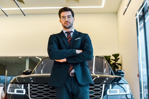 low angle view of businessman in eyeglasses posing with crossed arms near black automobile - Photo, Image