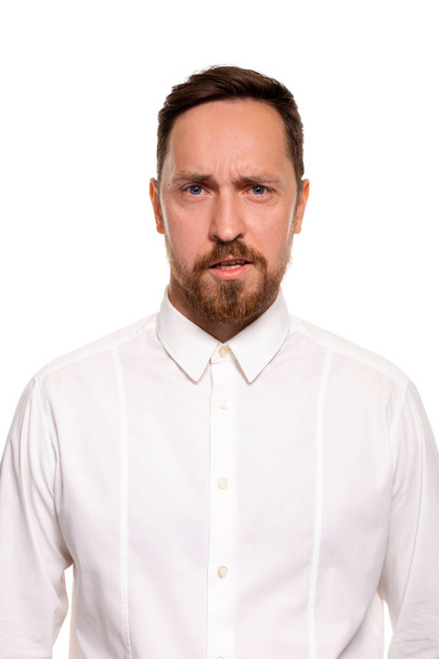 Retrato de hombre guapo con rastrojo tiene expresión indignante, frunce el ceño, vestido con camisa blanca, aislado sobre fondo blanco
. - Foto, imagen