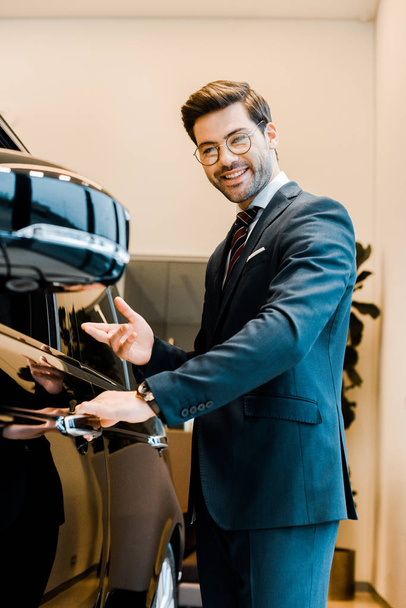 cheerful male car dealer in eyeglasses pointing at black automobile in car salon - Foto, imagen