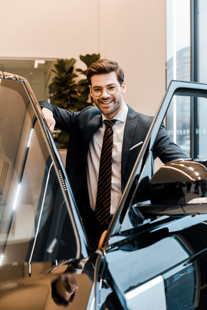 hombre de negocios riendo en anteojos posando cerca de automóvil en salón de coches
 - Foto, Imagen