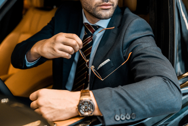 vista parziale dell'uomo d'affari con orologio di lusso che tiene gli occhiali mentre è seduto in auto
 - Foto, immagini