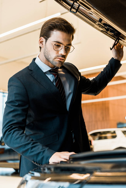low angle view of businessman in eyeglasses looking inside opened car hood of black automobile - Photo, Image