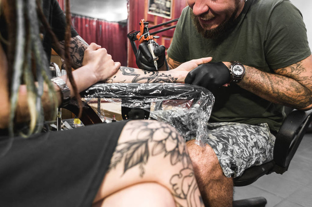 lány a raszta a egy tetováló szalonnal. A mester egy képet hoz létre, a test egy fiatal gyönyörű lány. Közeli kép a kéz és a tetováló gép - Fotó, kép