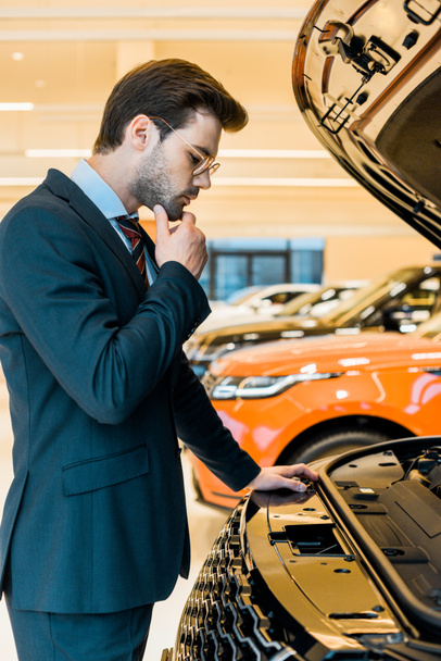 side view of thoughtful businessman looking at opened car hood of black automobile - Photo, Image