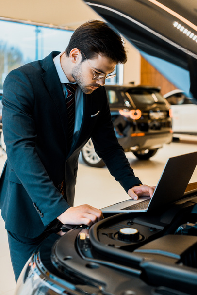 ориентированный молодой бизнесмен в очках проверяет капот автомобиля с ноутбуком
  - Фото, изображение