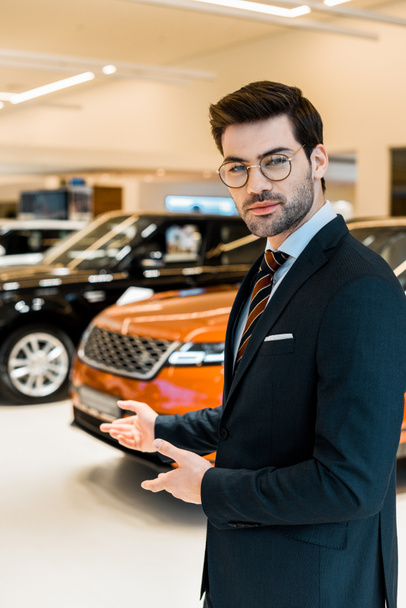 young male car dealer in eyeglasses pointing by hands in car salon - Foto, imagen