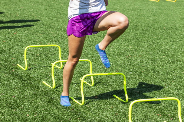 Un corredor de atletismo y atletismo no está usando zapatos mientras realiza ejercicios de forma pisando los mini obstáculos amarillos en un campo de césped en calcetines y pantalones cortos
. - Foto, imagen