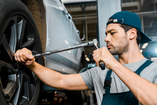 focused male auto mechanic in working overall working with wheel wrench at auto mechanic shop - Zdjęcie, obraz