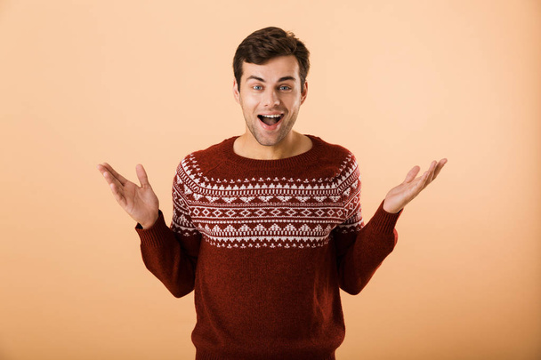 Image of joyful man 20s with bristle wearing knitted sweater laughing on camera with hands aside isolated over beige background - Φωτογραφία, εικόνα
