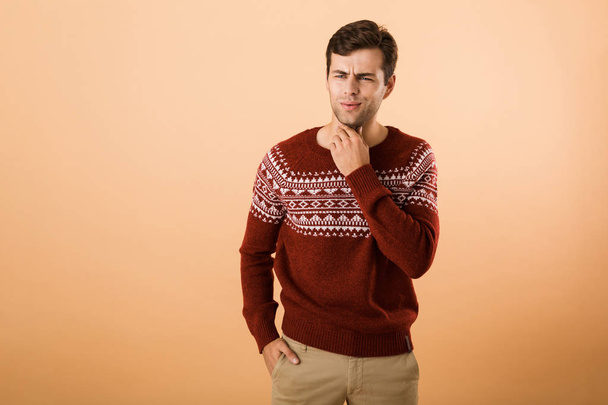 Image of surprised man 20s with bristle wearing knitted sweater looking aside and touching chin isolated over beige background - Фото, изображение