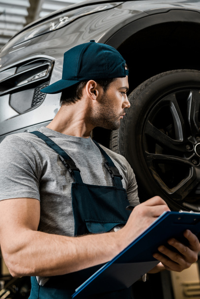 reparador concentrado en uniforme con el bloc de notas comprobar las ruedas del coche en el taller de reparación de automóviles
 - Foto, imagen