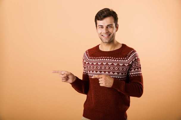 Image of brunette man 20s with bristle wearing knitted sweater pointing finger aside at copyspace isolated over beige background - Foto, imagen