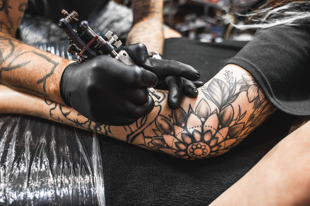 girl with dreadlocks in a tattoo parlor. The master creates a picture on the body of a young beautiful girl. Close-up of hands and tattoo machine - Zdjęcie, obraz