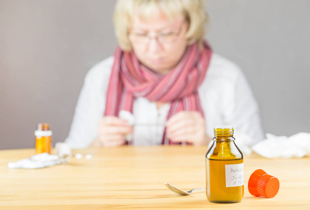 Uma garrafa com a inscrição alemã para xarope de tosse, no fundo fora de foco uma mulher com cachecol olhando para um termômetro febre
 - Foto, Imagem
