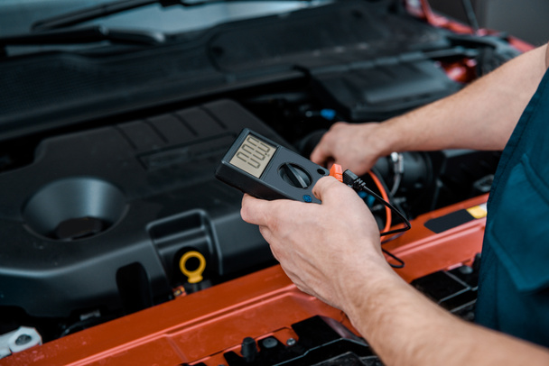 Teilansicht des Automechanikers mit Multimeter-Voltmeter zur Überprüfung der Autobatteriespannung im Mechaniker-Geschäft - Foto, Bild