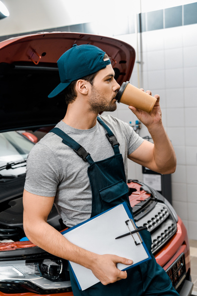 young mechanic with notepad drinking coffee and leaning on car with opened cowl at mechanic shop - Foto, imagen