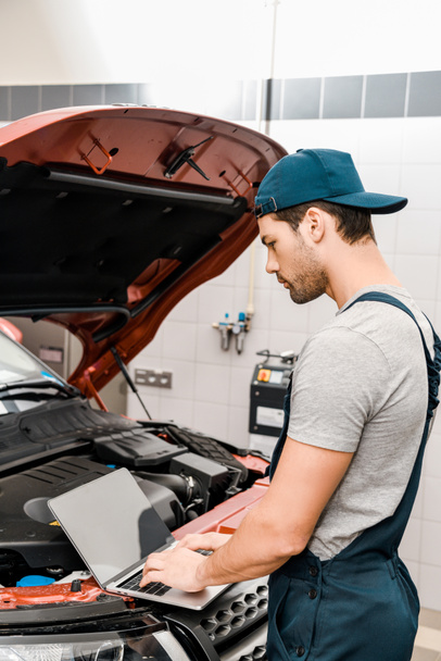 side view of auto mechanic working on laptop at automobile with opened car cowl at mechanic shop - Φωτογραφία, εικόνα
