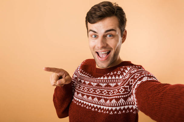 Image of young man 20s with bristle wearing knitted sweater pointing finger aside while taking selfie photo isolated over beige background - Fotoğraf, Görsel