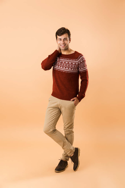 Full length image of brunette man 20s with bristle wearing knitted sweater smiling isolated over beige background - Fotoğraf, Görsel