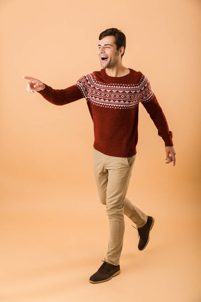 Full length image of happy man 20s with bristle wearing knitted sweater smiling while walking isolated over beige background - Фото, изображение