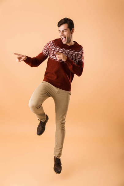 Full length image of caucasian man 20s with bristle wearing knitted sweater laughing and pointing aside isolated over beige background - Foto, imagen