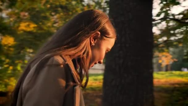 Motion footage, gorgeous caucasian girl in profile mowing and walking near camera in golden fall park and happily enjoying sunlights on her face, natural artistic illustration outside - Imágenes, Vídeo