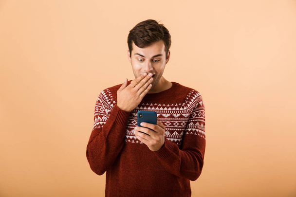 Image of surprised man 20s with stubble wearing knitted sweater using cell phone isolated over beige background - Φωτογραφία, εικόνα