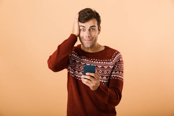 Image of joyous man 20s with stubble wearing knitted sweater using mobile phone isolated over beige background - Foto, Imagem