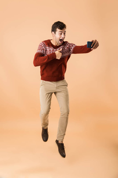 Full length portrait a cheerful young man jumping isolated over beige background, taking selfie with mobile phone, showing thumbs up - Foto, Bild