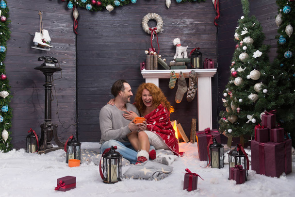 Эмоциональная любящая пара обнимается у камина в новогоднем интерьере и смеется, когда пьет чай
 - Фото, изображение